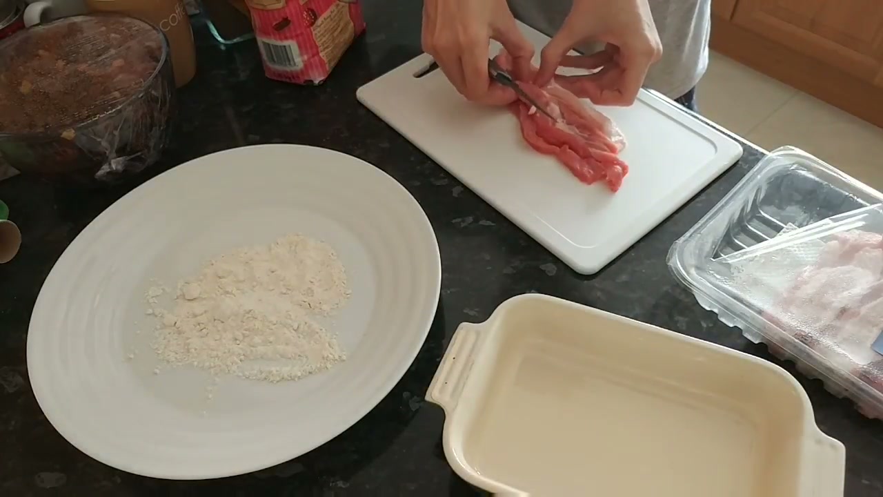 Hands preparing meat on a cutting board beside a plate of coating ingredients and a rectangular dish.