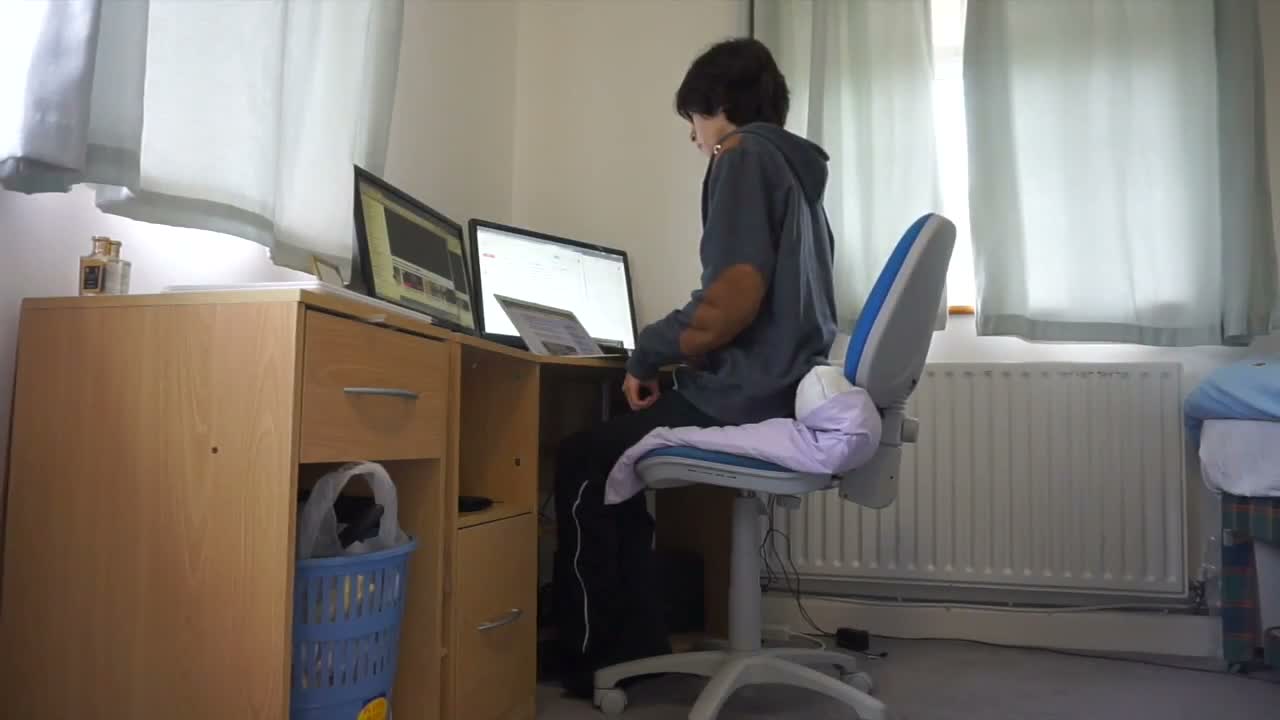 An on-camera HIMR participant seated at a desk with two computer displays in a bedroom-like room