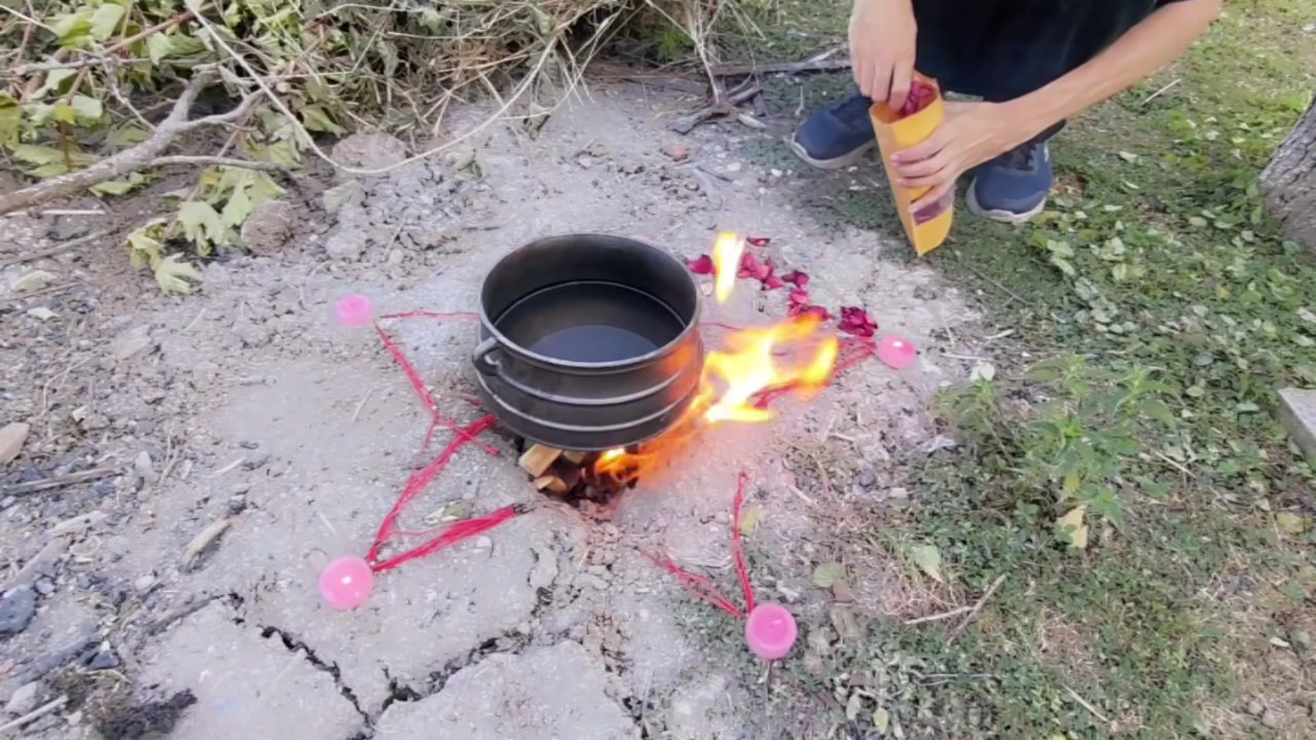 Black pot above a small outdoor fire, surrounded by a red five-pointed design, pink candles, and rose petals