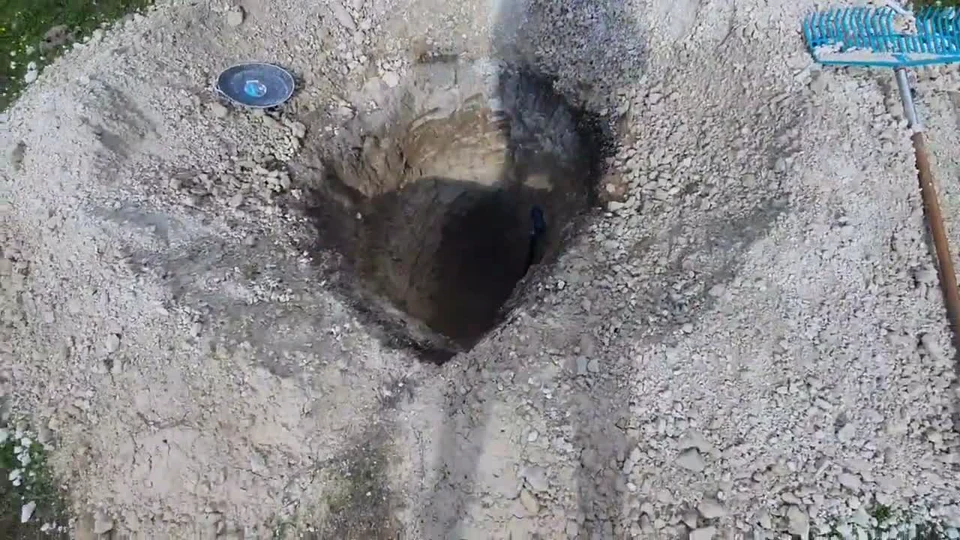 A large excavation in earth with displaced soil, a shallow container, and a blue rake visible nearby