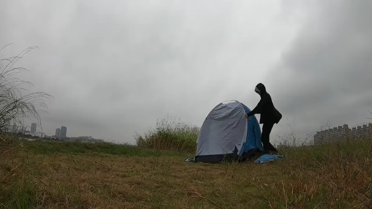 Daniel standing beside a blue tent in a grassy riverside area under an overcast sky