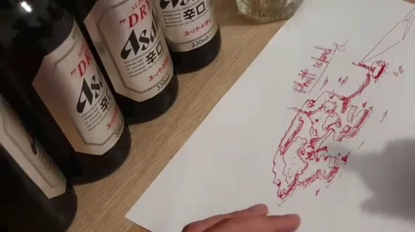 A red-ink island drawing on paper, with a hand at the lower edge and bottles beside the sheet.
