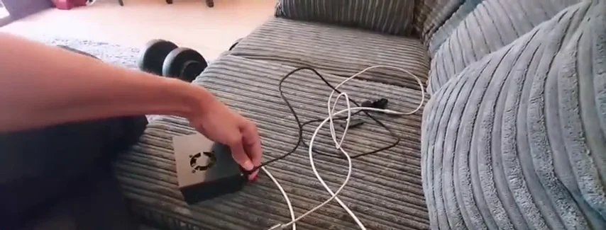 A hand handling a small fan-equipped box beside connected black and white cables on a sofa.